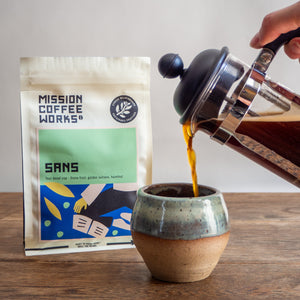 Person pouring coffee from a French press into a ceramic cup with Mission Coffee Works package in the background.