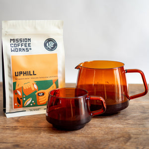 Mission Coffee Works coffee package with an amber glass mug and pourer on a wooden surface